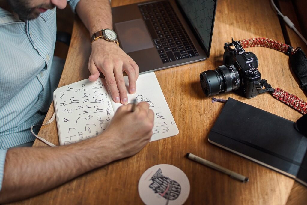 A man sketching hand-lettered notes on paper at a desk with a laptop and camera nearby