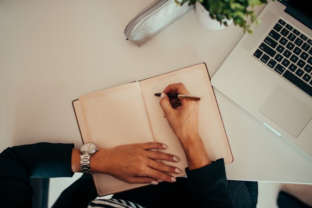 A person writing notes in an open notebook at a desk beside an open laptop