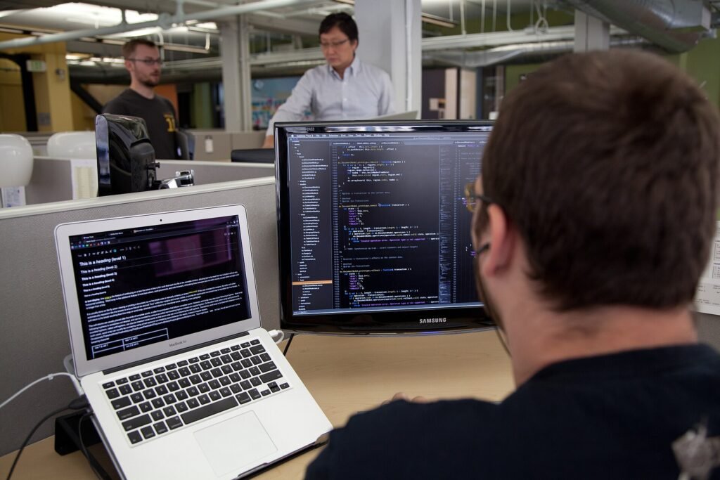 A software engineer working at a desk with code on a laptop and a large monitor in an open-plan office