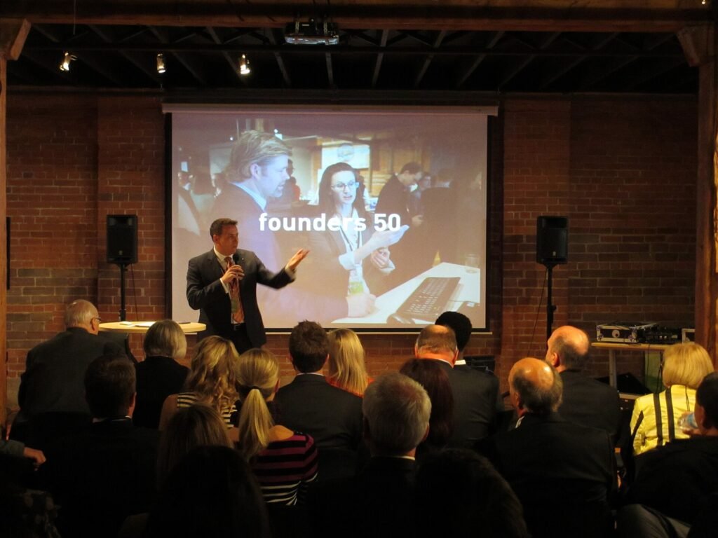A speaker presenting to a seated audience in a brick-walled venue under a "founders 50" slide