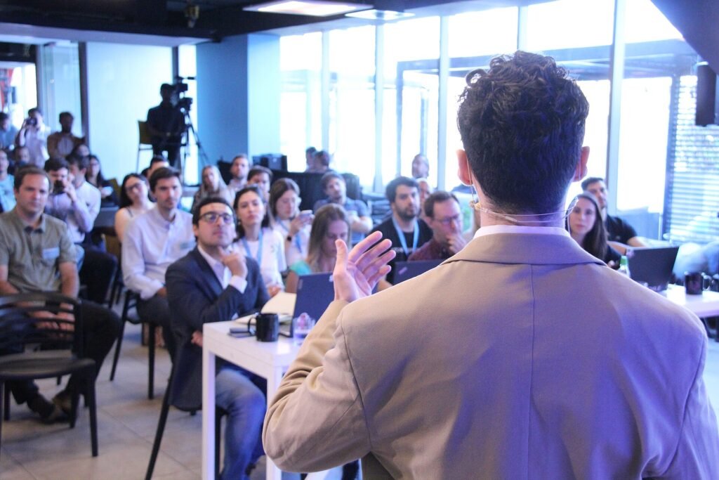 A speaker seen from behind addressing a seated audience at a startup demo day