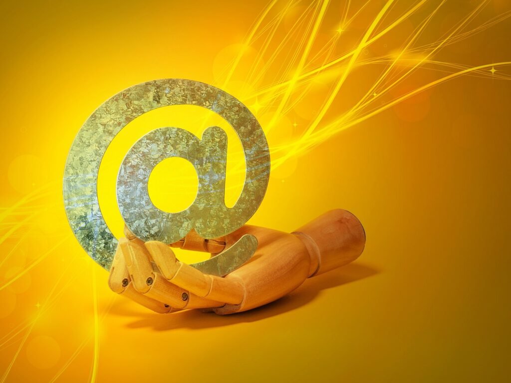 A wooden mannequin hand holding a large metallic @ symbol against a yellow background