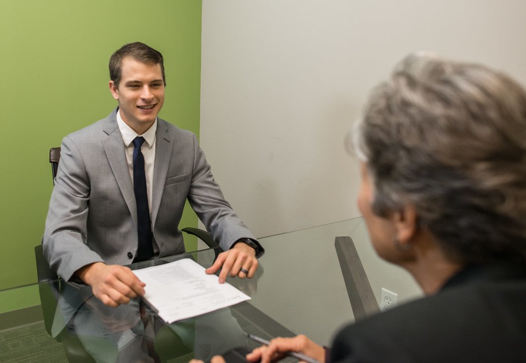 A young man in a shirt and tie sitting across a desk from an interviewer