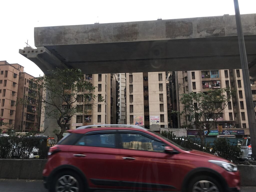 An elevated metro viaduct under construction over a road in Mumbai, with apartment towers behind