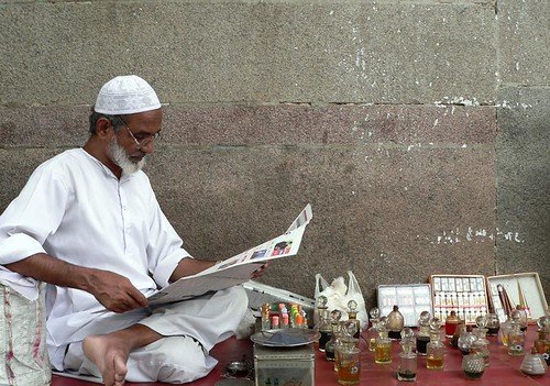 An itr (attar) seller in a white cap reading a newspaper beside rows of small glass perfume bottles