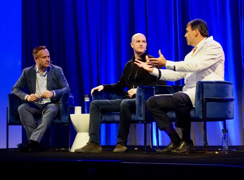 Brian Armstrong seated on stage beside investor Tim Draper, who is gesturing as he speaks, in front of a blue curtain