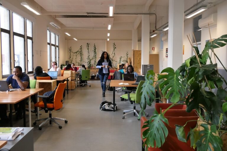 Open-plan coworking space with people working at shared desks under large windows
