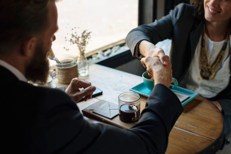 Two business people shaking hands across a cafe table with coffee and a phone